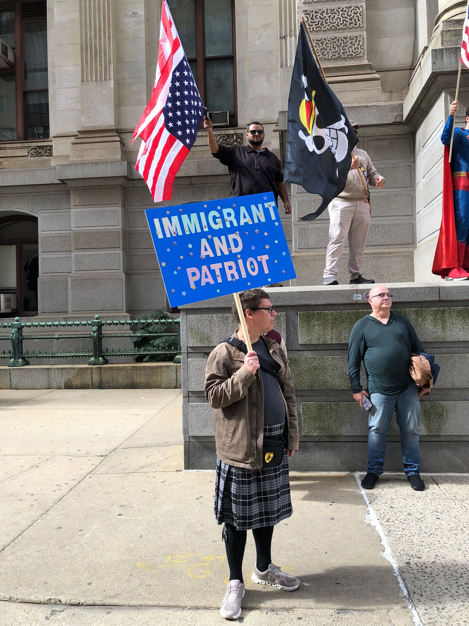 A young white man in a jacket and kilt holds a spangled blue sign that says “immigrant and patriot”