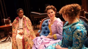 Three women dressed in colonial-style clothing next to each other, two of them looking at the one in the center, incredulous