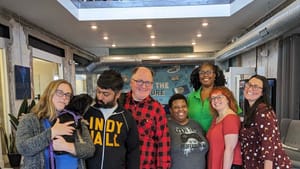 A multi-racial group of smiling colleagues posing together in a lofty coworking space. Alaina holds a tiny dog on the left.