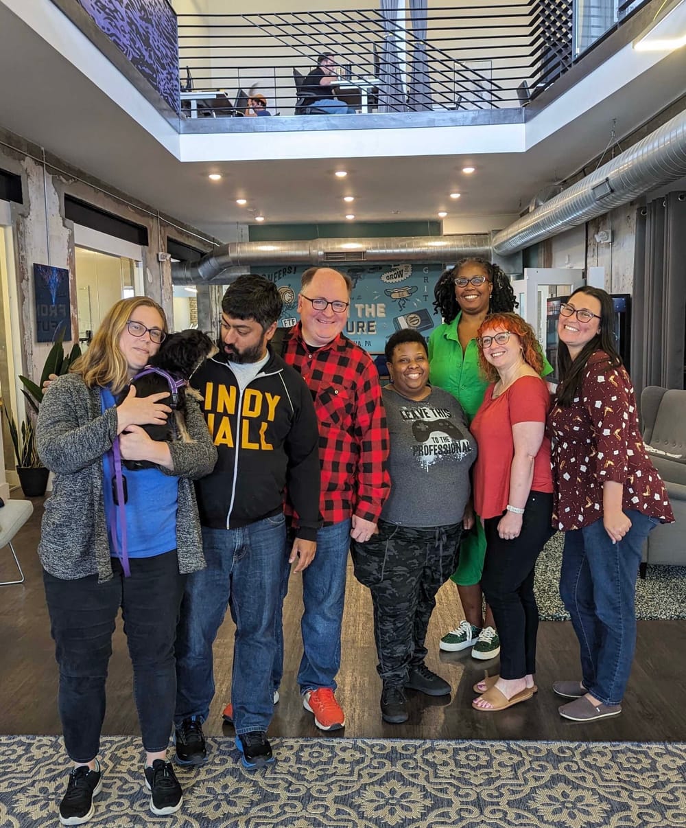 A multi-racial group of smiling colleagues posing together in a lofty coworking space. Alaina holds a tiny dog on the left.