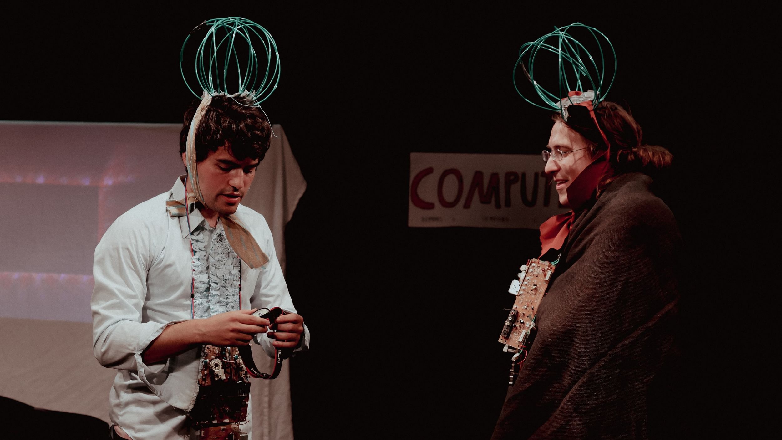 Two people on stage, both with wire coils on their heads, fiddle with computer parts