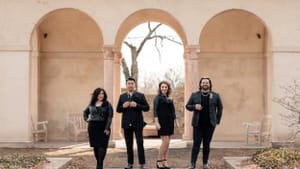 Portrait, four people in black formalwear in an outdoor area with a series of arches part of a larger building is behind them