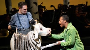 Kanagawa, in denim shirt and suspenders, manipulates an emotive cow puppet, which Thompson, in a green jacket, sings to.