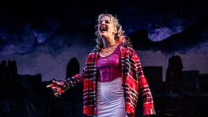 O’Hare, a white woman with curly blond hair, yells defiantly. She wears a white skirt, purple bodice and red plaid shirt.