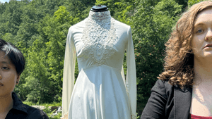 The couple stands solemnly facing forward, wearing black, hands joined in front of a Victorian wedding dress on a mannequin.