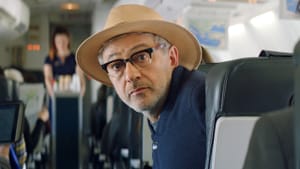 A man in a wide round-brimmed hat and black framed glasses looks back from his seat on an airplane, inquisitive