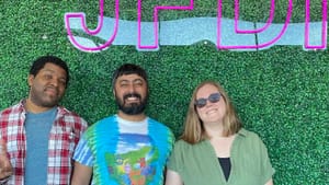 Kyle, Neil, and Alaina line up smiling against a faux green backdrop with pink letters spelling JFDI.