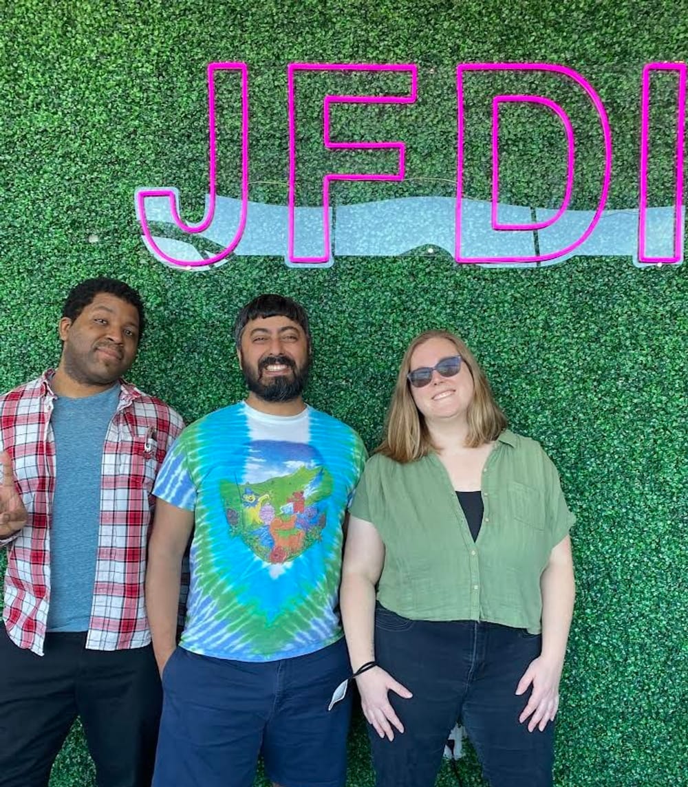 Kyle, Neil, and Alaina line up smiling against a faux green backdrop with pink letters spelling JFDI.