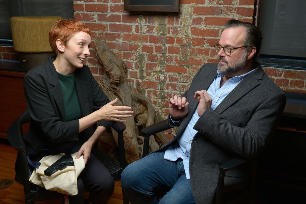 Two performers sit in a brick-walled room, one has his hands up in reservation, the other open handed looking at him