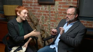 Two performers sit in a brick-walled room, one has his hands up in reservation, the other open handed looking at him