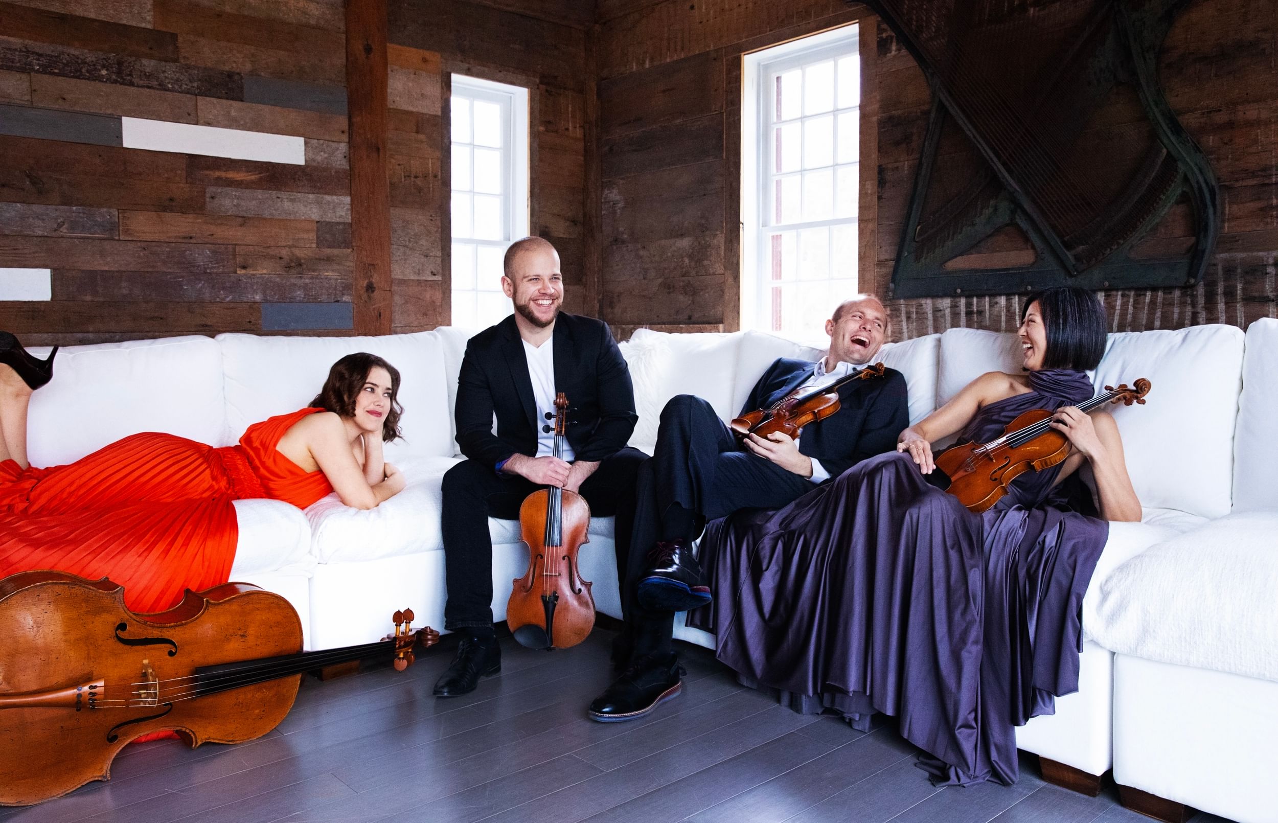 Four players laughing on a long white couch. Three hold violins, a cello on the floor, its front facing the camera