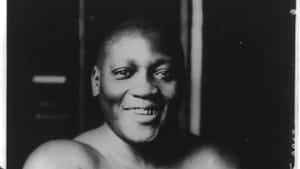 A black and white portrait of Jack Johnson, his arms crossed, smiling. He’s topless, wearing a white towel or pair of shorts.