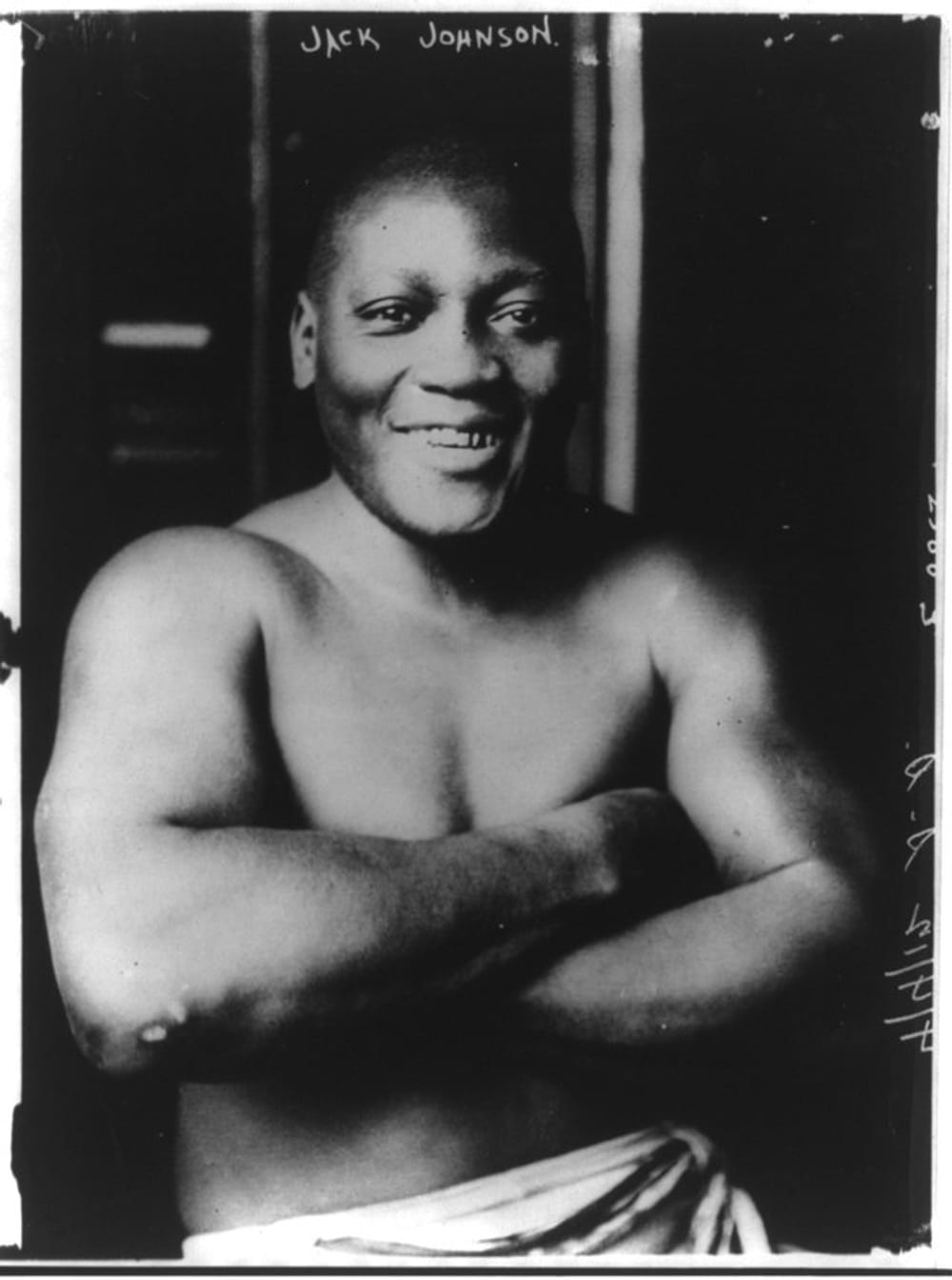 A black and white portrait of Jack Johnson, his arms crossed, smiling. He’s topless, wearing a white towel or pair of shorts.