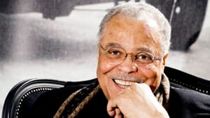 Sitting in a large black chair, wearing a black suit and brown scarf, Jones smiles with one hand touching his chin.