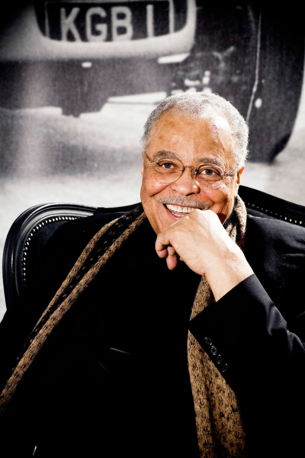 Sitting in a large black chair, wearing a black suit and brown scarf, Jones smiles with one hand touching his chin.