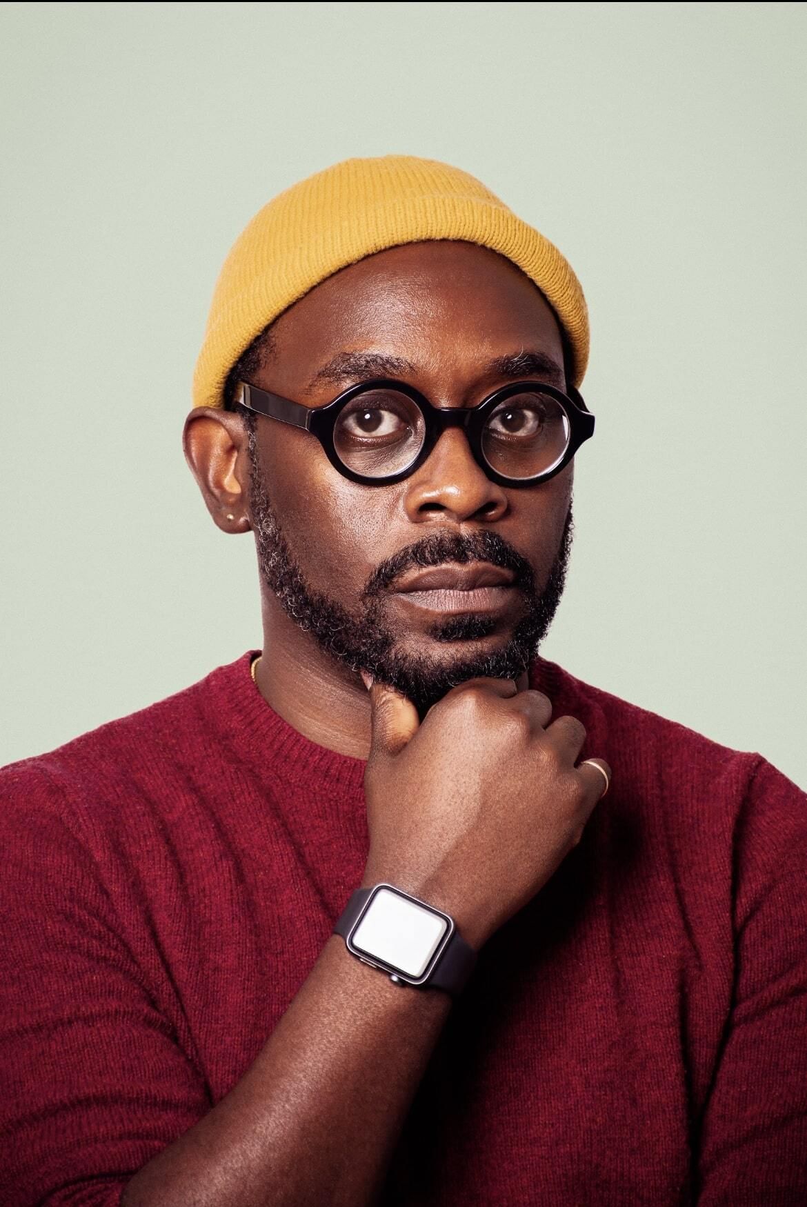 Headshot of Ijames, a Black man wearing round glasses and a yellow beanie. He thoughtfully puts his hand to his chin.