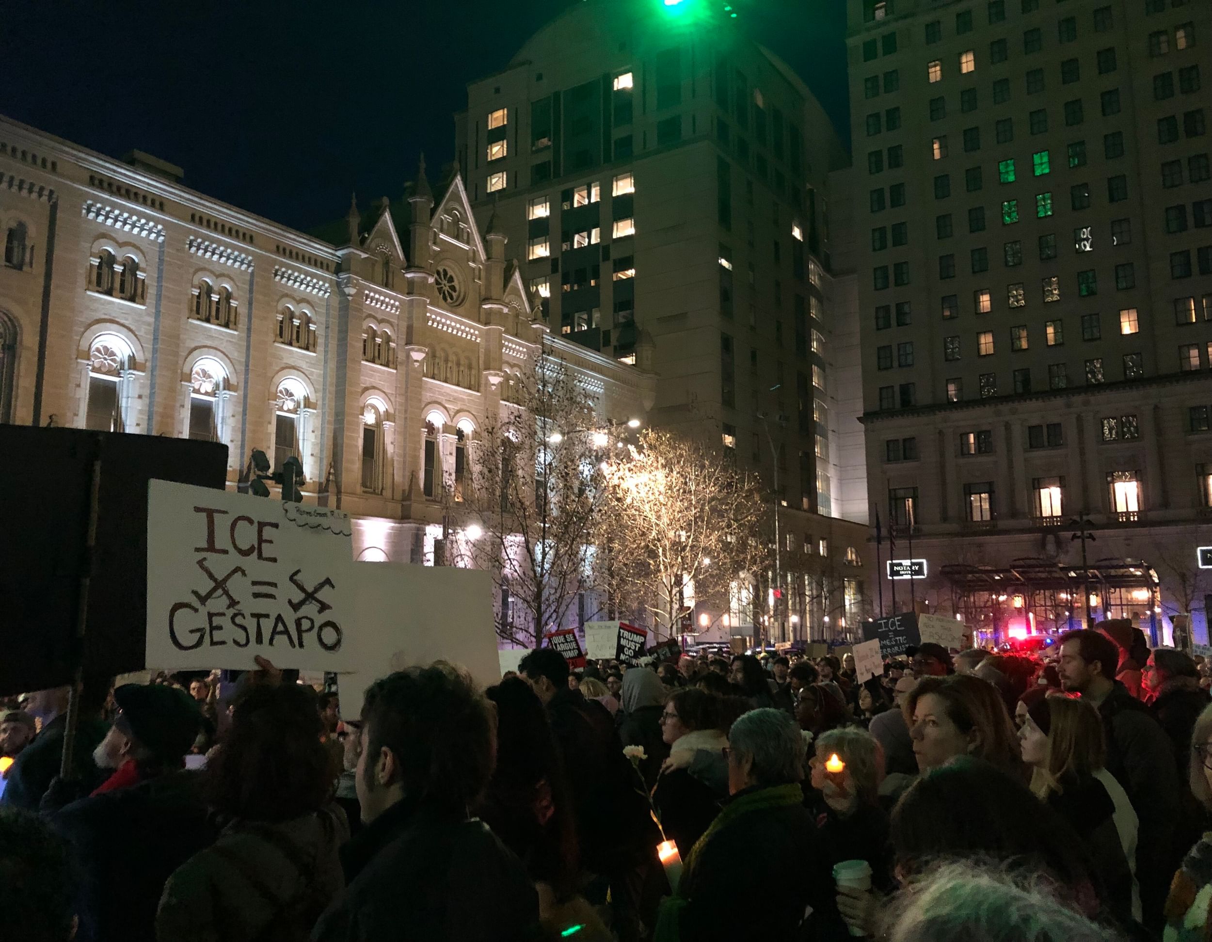 Nighttime protest shows a crowd of hundreds, with a sign comparing ICE to the Nazi gestapo in the foreground.