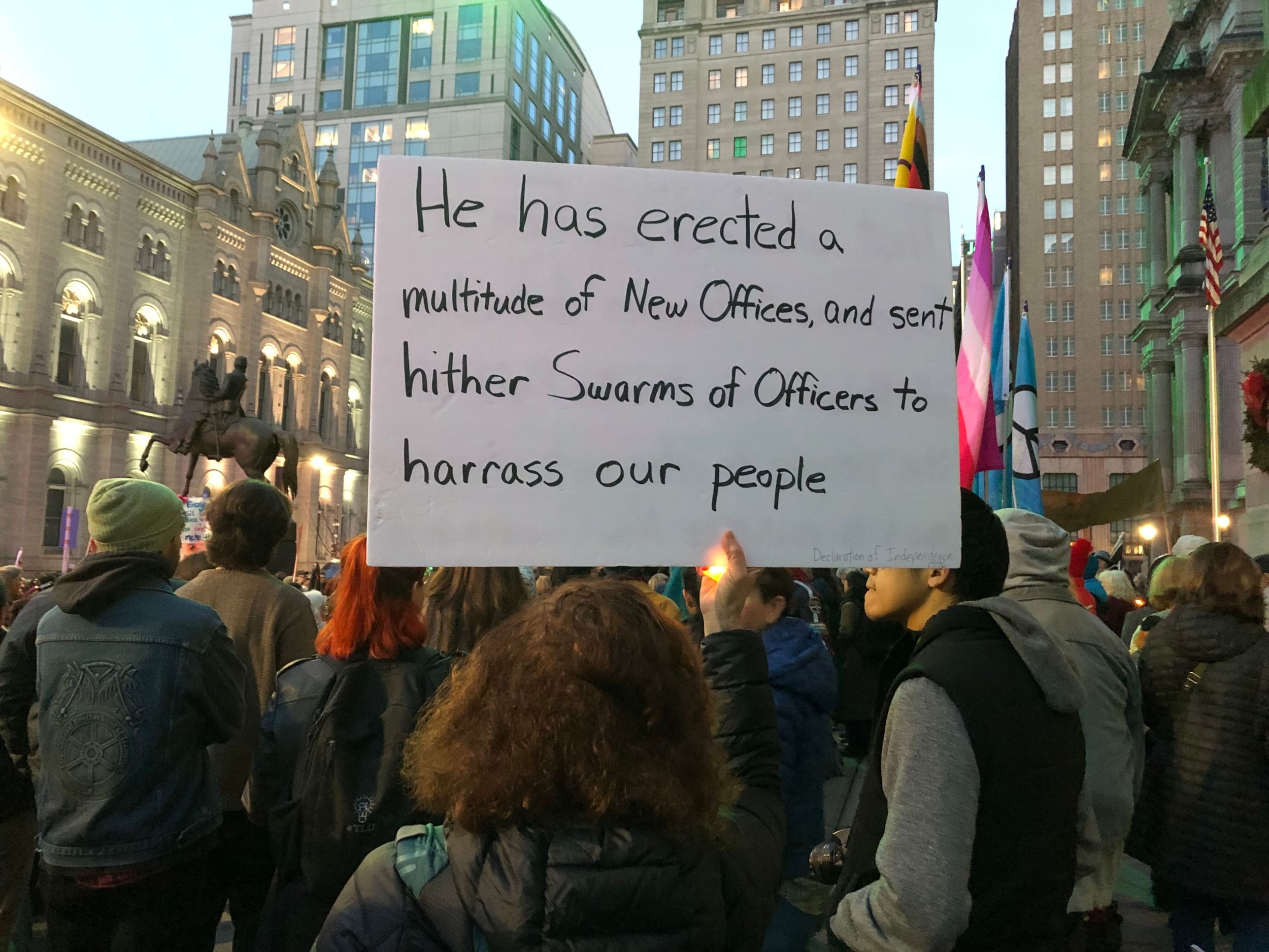 Crowd of protestors seen from behind, including a sign quoting the Declaration about the king's officers harassing people