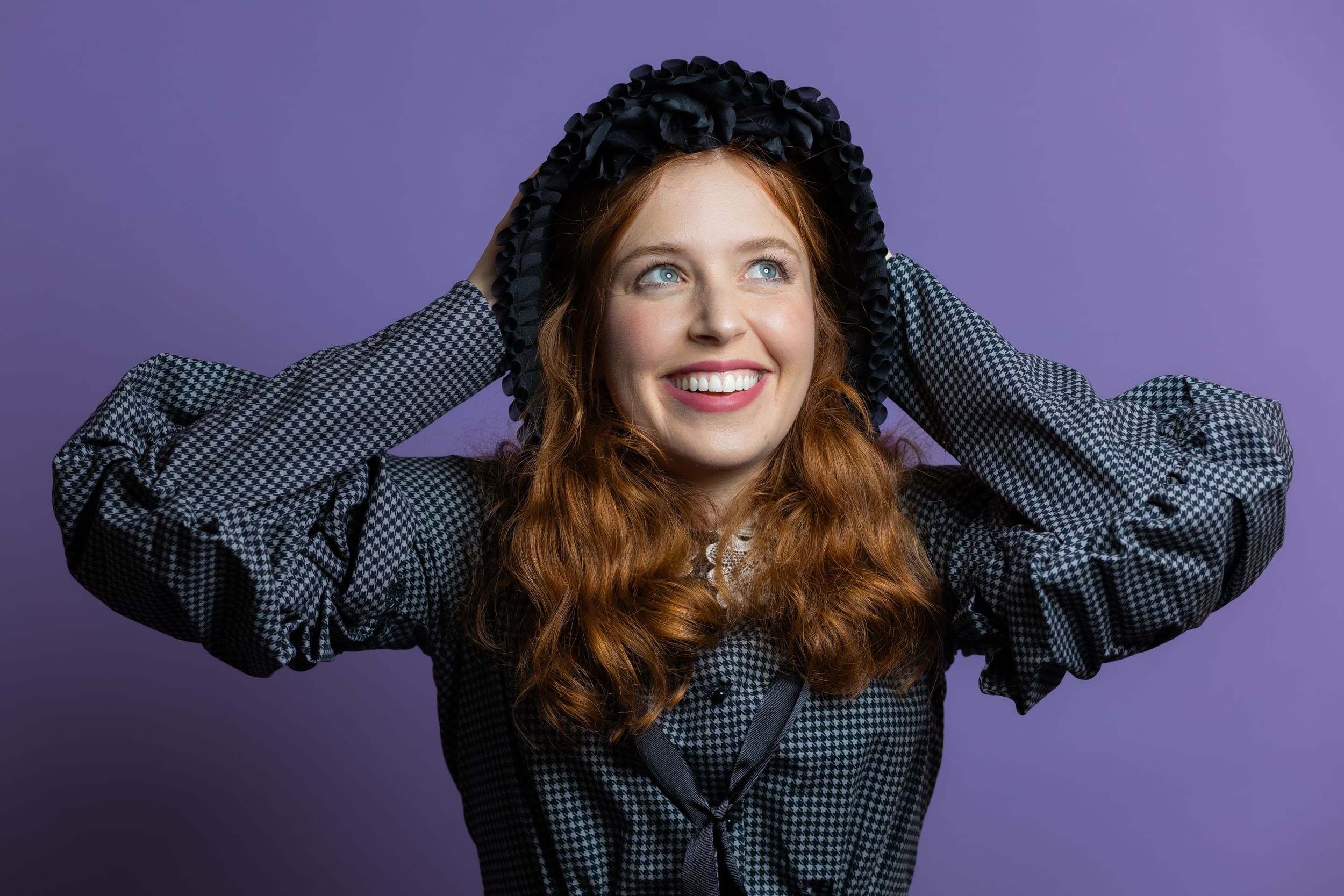 Polistina, a young white woman with wavy red hair and 19th-century garb, smiles with her hands on her head.