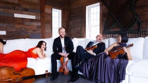 A group of musicians, dressed up, in laughter, spread out over a long, wide white couch, bright windows behind them