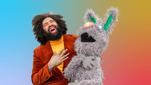 Jayson Musson, a Black man in an orange suit, holds the furry gray puppet on his lap. They both tip their heads back laughing