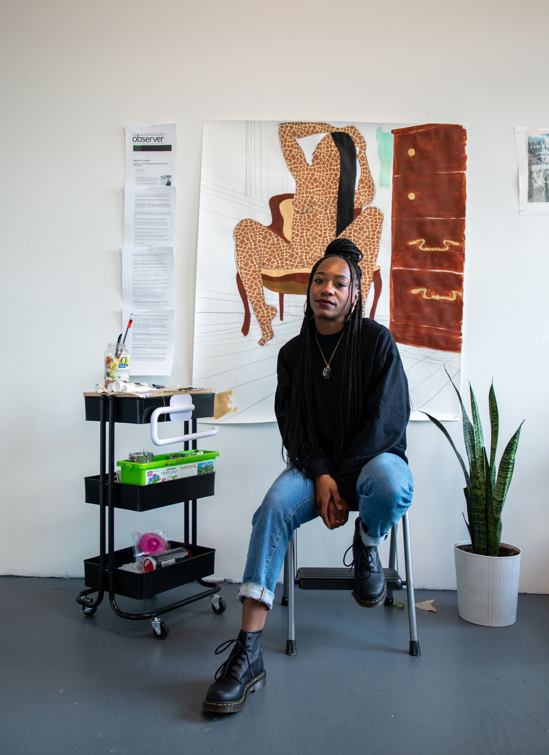 Sabree, a Black woman, sits in front of one of her works in what looks like a minimalistic studio space