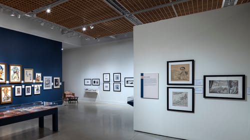 White and dark blue gallery walls hold many framed works with a vitrine at center holding period publications.