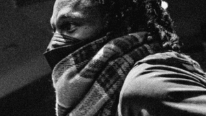 Black & white photo of Page, a Black man wearing a facemask and a plaid scarf, seen in profile.