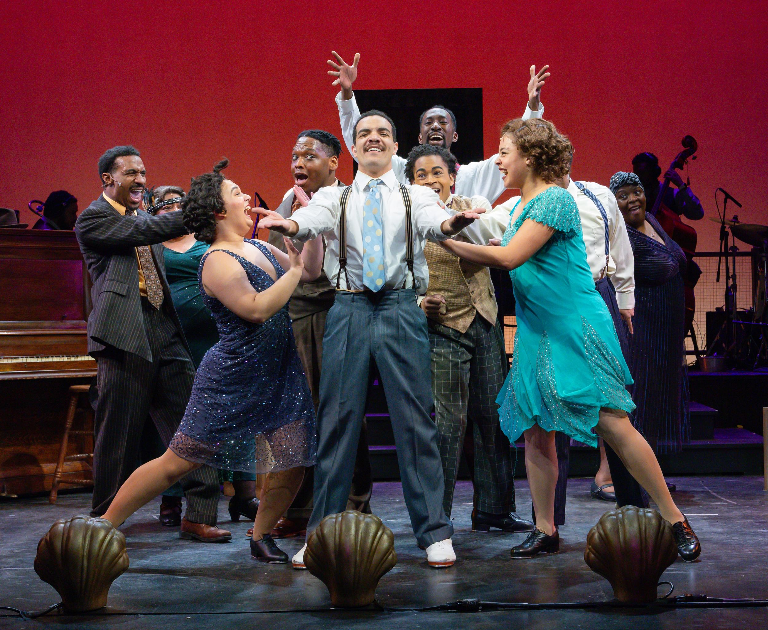 Martin, in tie and suspenders, stretches his hands forward as nine cast members pose ecstatically around him.