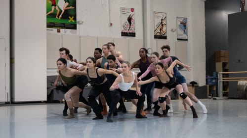 Multiracial group of 14 dancers in rehearsal gear, leaning forward intensely as one in the BalletX studio.