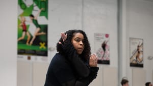 Archibald, a Black woman in black leggings & sweatshirt, strikes an angled pose in the gray-floored BalletX studio.