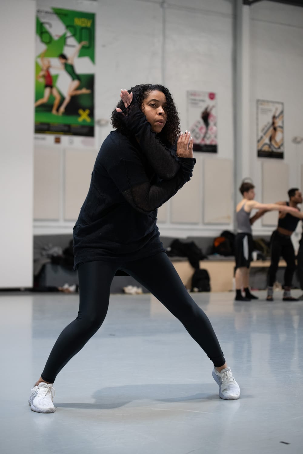 Archibald, a Black woman in black leggings & sweatshirt, strikes an angled pose in the gray-floored BalletX studio.