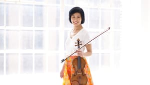 Koh, an Asian woman, in white shirt & orange floral skirt. She holds her violin & smiles against an airy white background