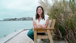 Weiner, a white woman with brown hair in jeans and a white tank, poses smiling on an outdoor bench overlooking a bay.