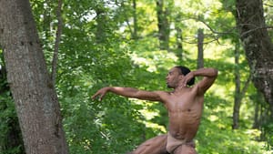 Dancer Joe Gonzalez dances outdoors, summertime trees behind him. He leaps so high he seems to be flying, arms & legs poised