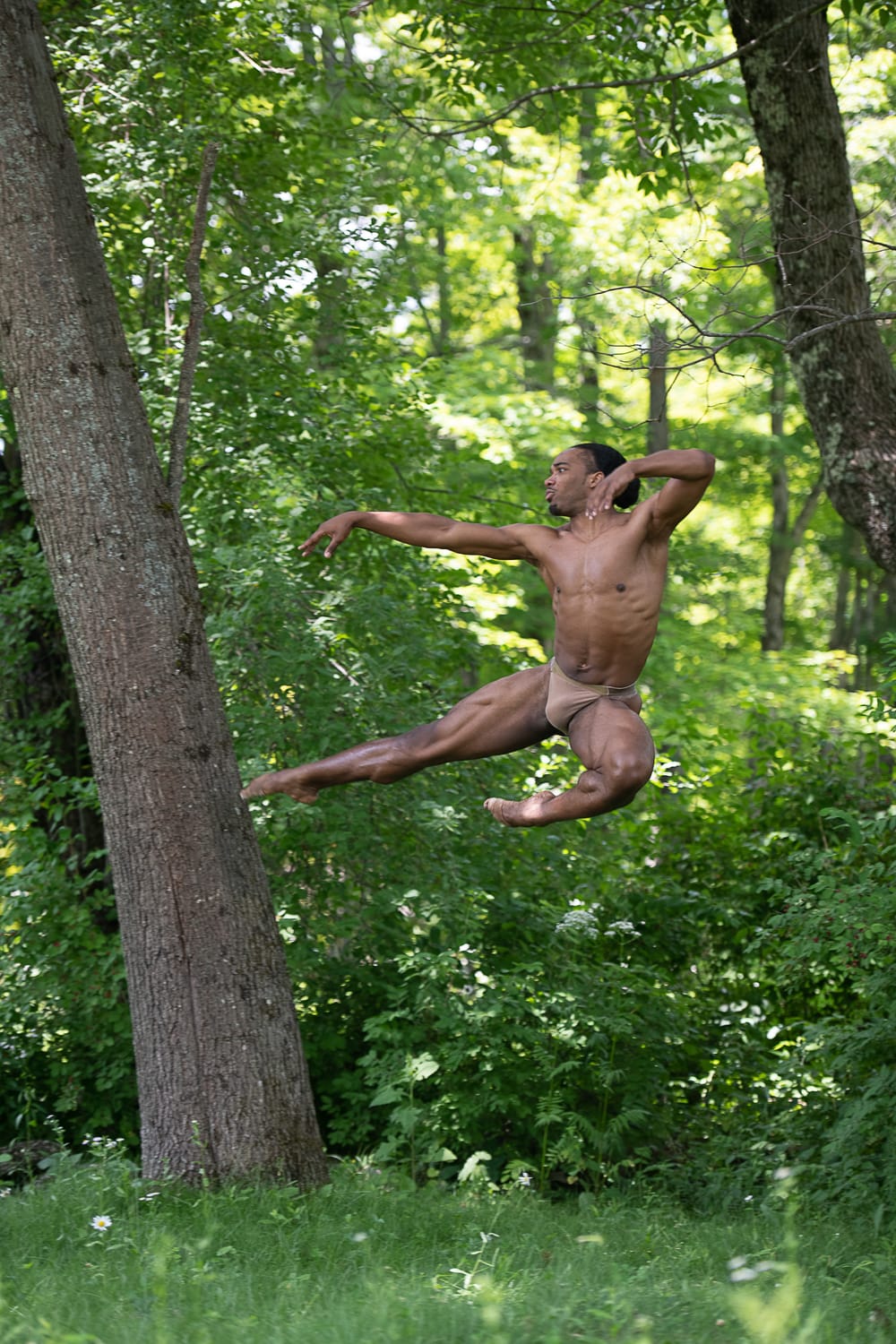 Dancer Joe Gonzalez dances outdoors, summertime trees behind him. He leaps so high he seems to be flying, arms & legs poised