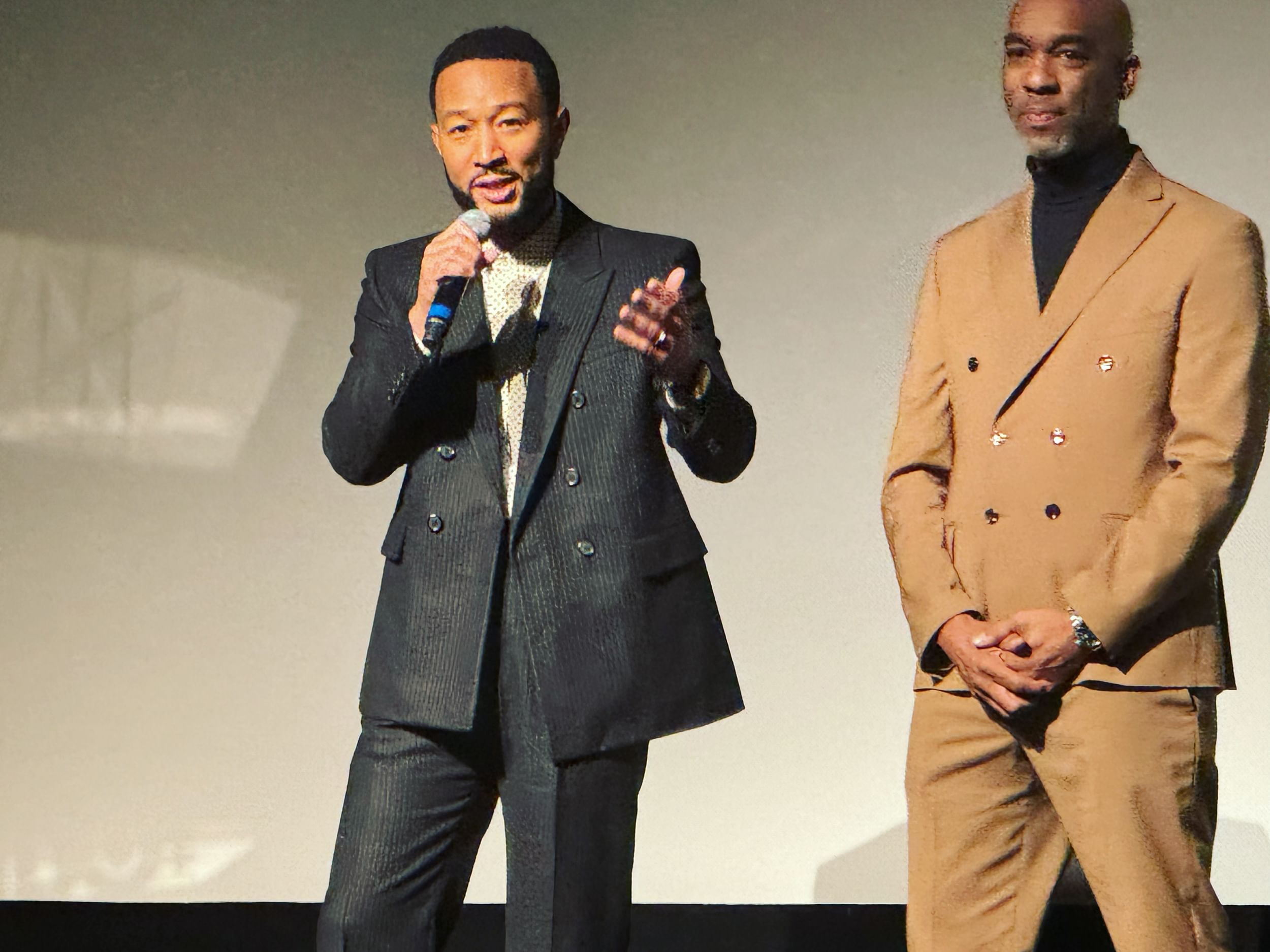 Legend, in a dark pinstripe suit, speaks into a microphone onstage. Jackson stands beside him in a light brown suit.