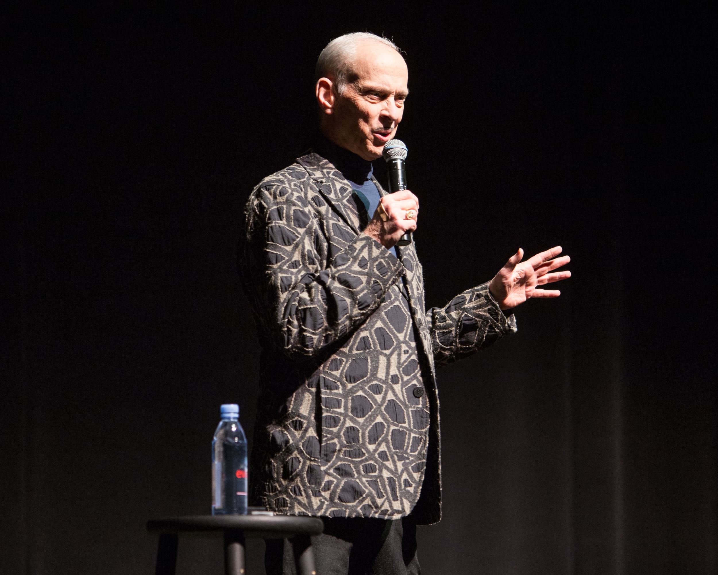 A man on stage in a blazer with a decorative design, holds a mic as he speaks out towards the audience