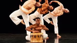 Three male dancers in white pants strike the same balletic pose bending to the left behind a fourth sitting & beating a drum