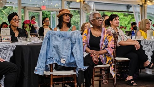 Close-up on about a dozen members of the multi-racial audience, sitting at tables in a lit-up tent at dusk.