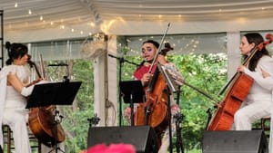 The women cellists wear white and de Jesús wears a tan bodysuit painted with blue and red, singing into a mic as they play