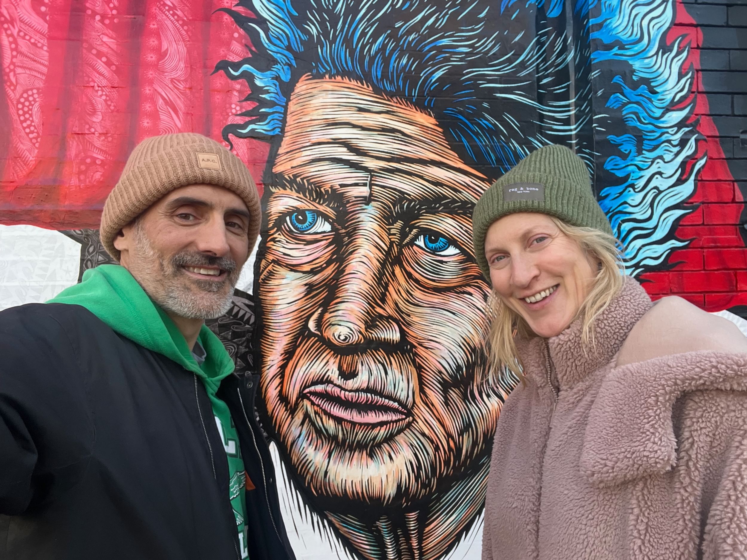 Suaudeau and Duchene in jackets and beanie caps smile for a group selfie in front of a colorful mural of David Lynch