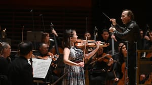 Kang, an Asian woman wearing a sleeveless patterned dress, plays the violin alongside the orchestra and conductor.
