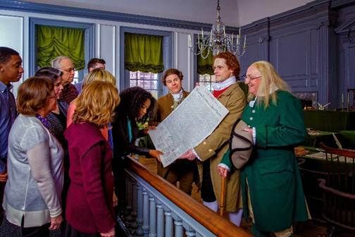 Inside the historic building, a group of visitors look up close at a copy of the Declaration held by a Jefferson re-enactor.