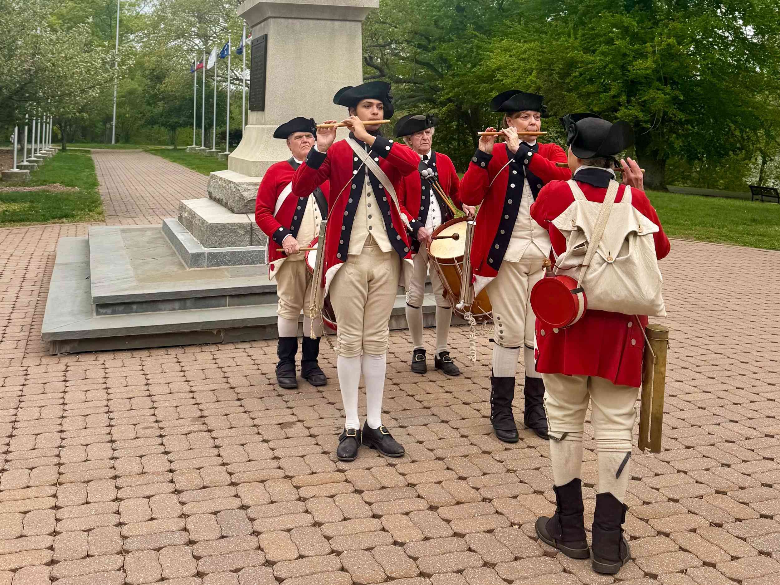 Five people dressed as Revolutionary soldiers play fifes and drums outdoors next to a stone monument.