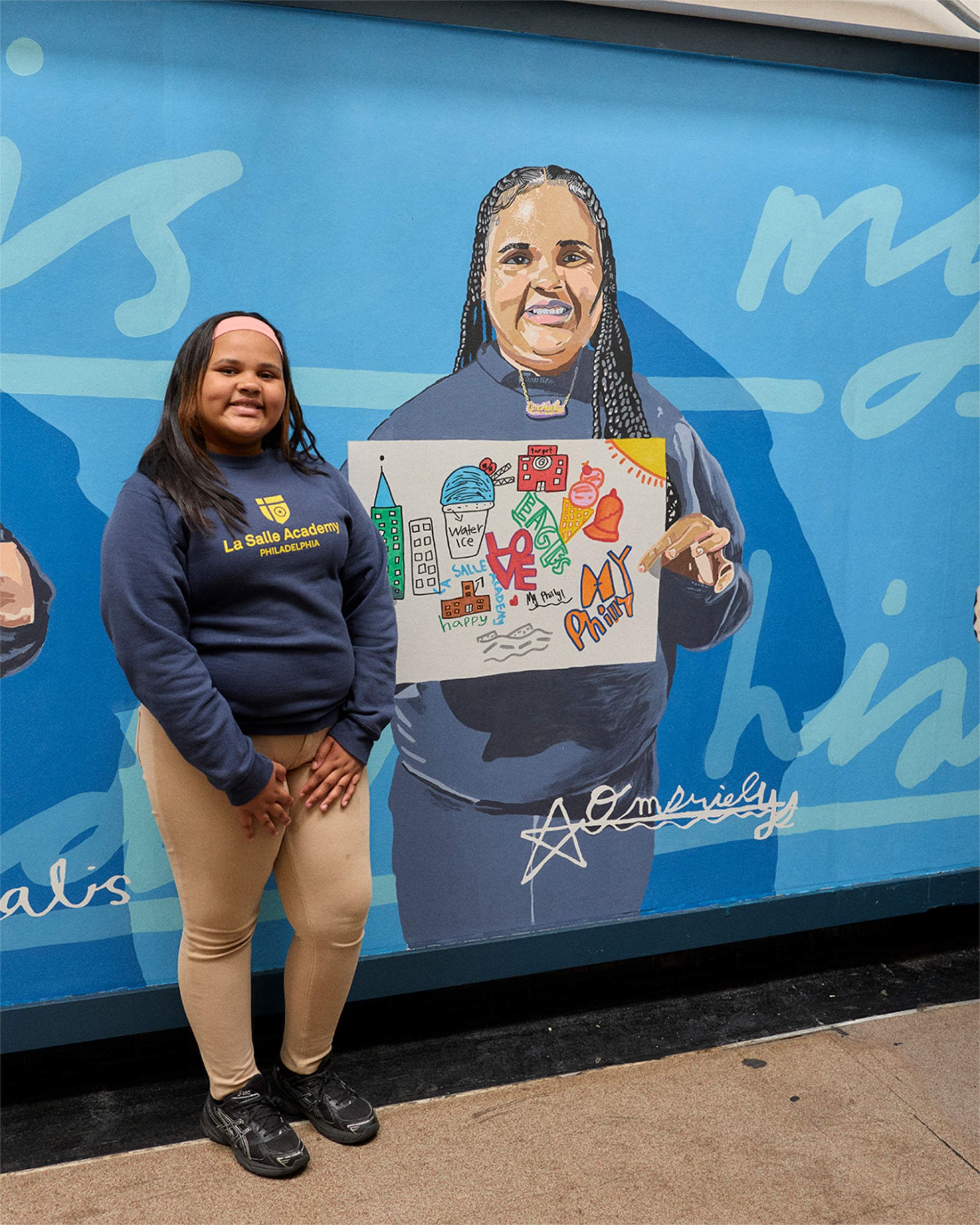 Closeup on a smiling elementary schooler in school sweatshirt posting next to a large portrait of themselves holding artwork