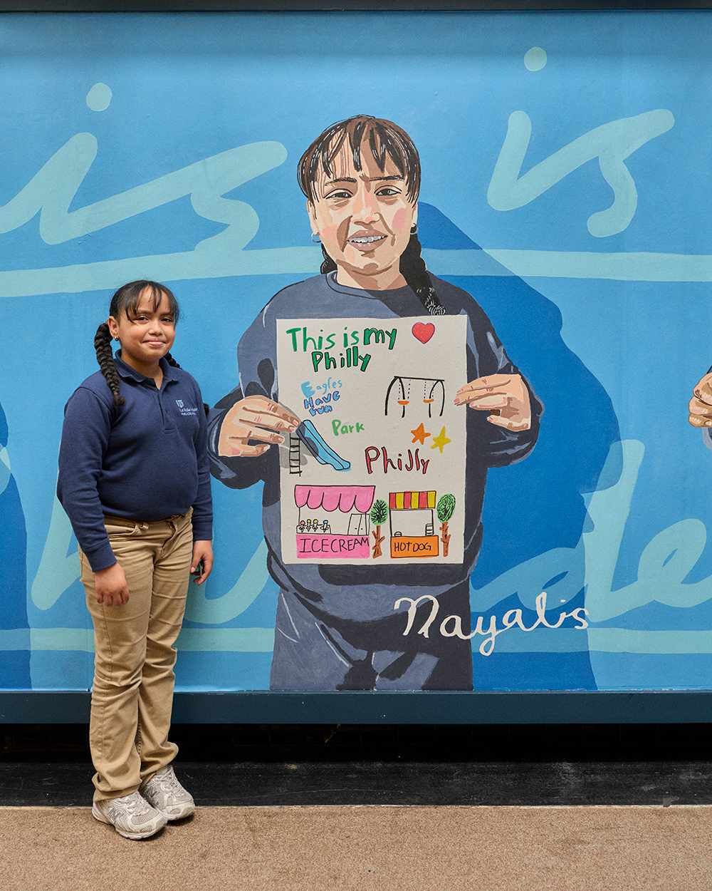 Closeup on a smiling elementary schooler in school sweatshirt posting next to a large portrait of themselves holding artwork