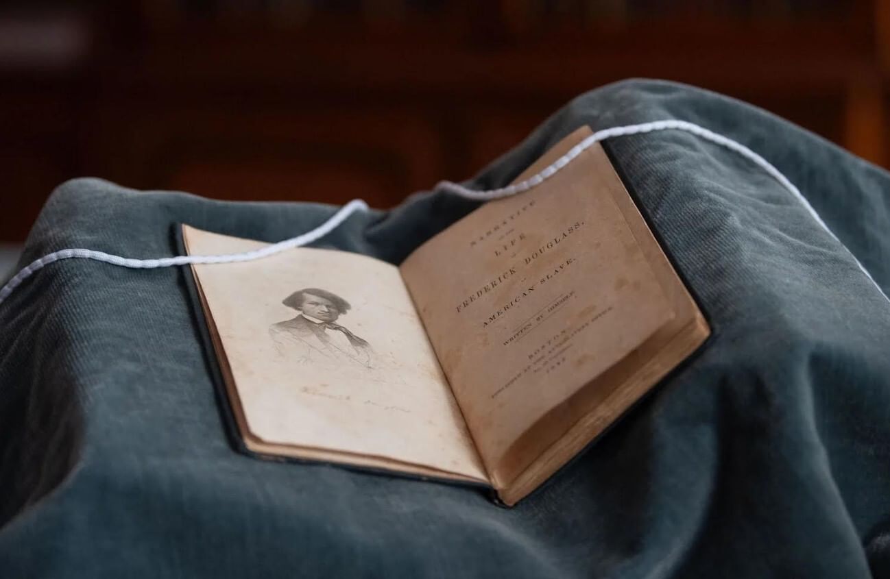 Closeup photo on the small historic book open to its title page with a portrait of Douglass, resting on soft gray fabric.