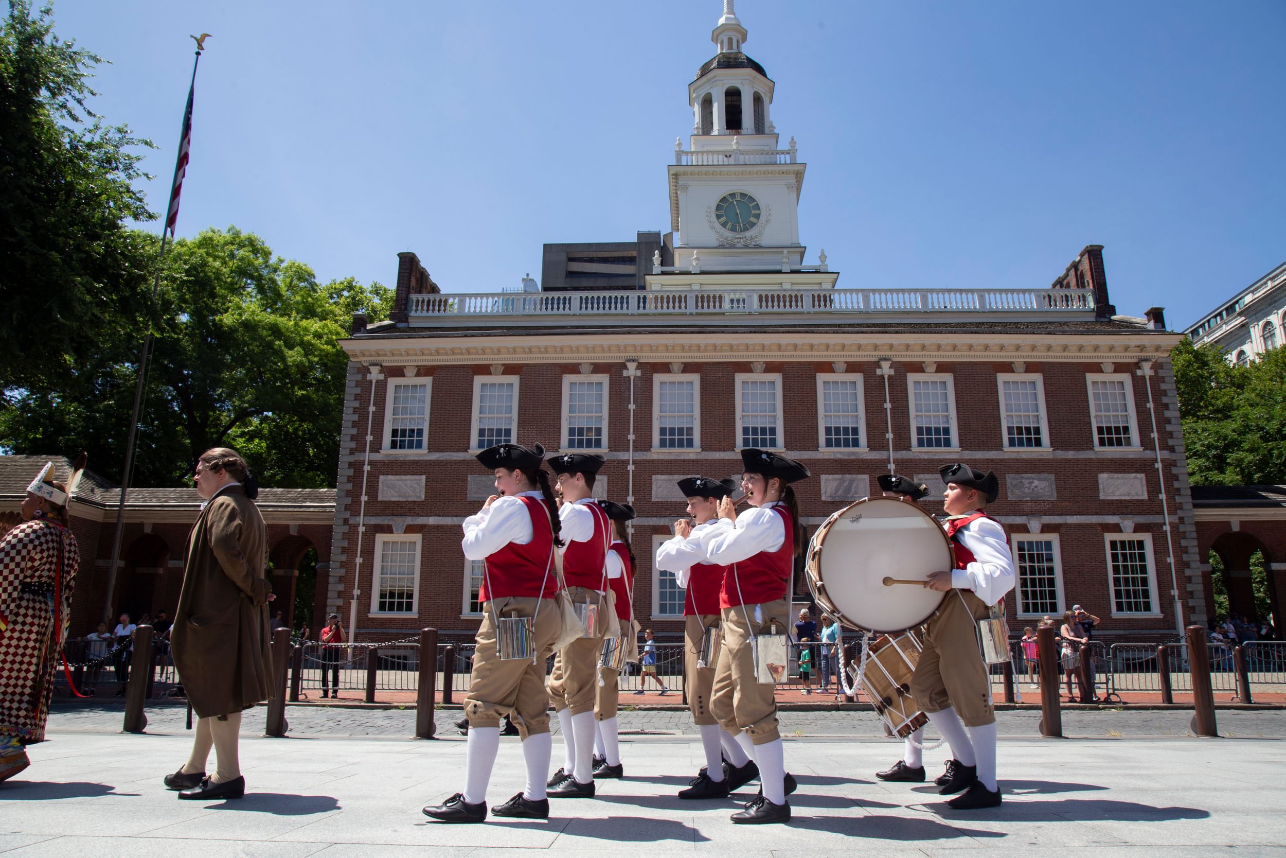 A troupe of about 7 kids in Colonial garb march while playing fifes and drums past the brick façade of Independence Hall