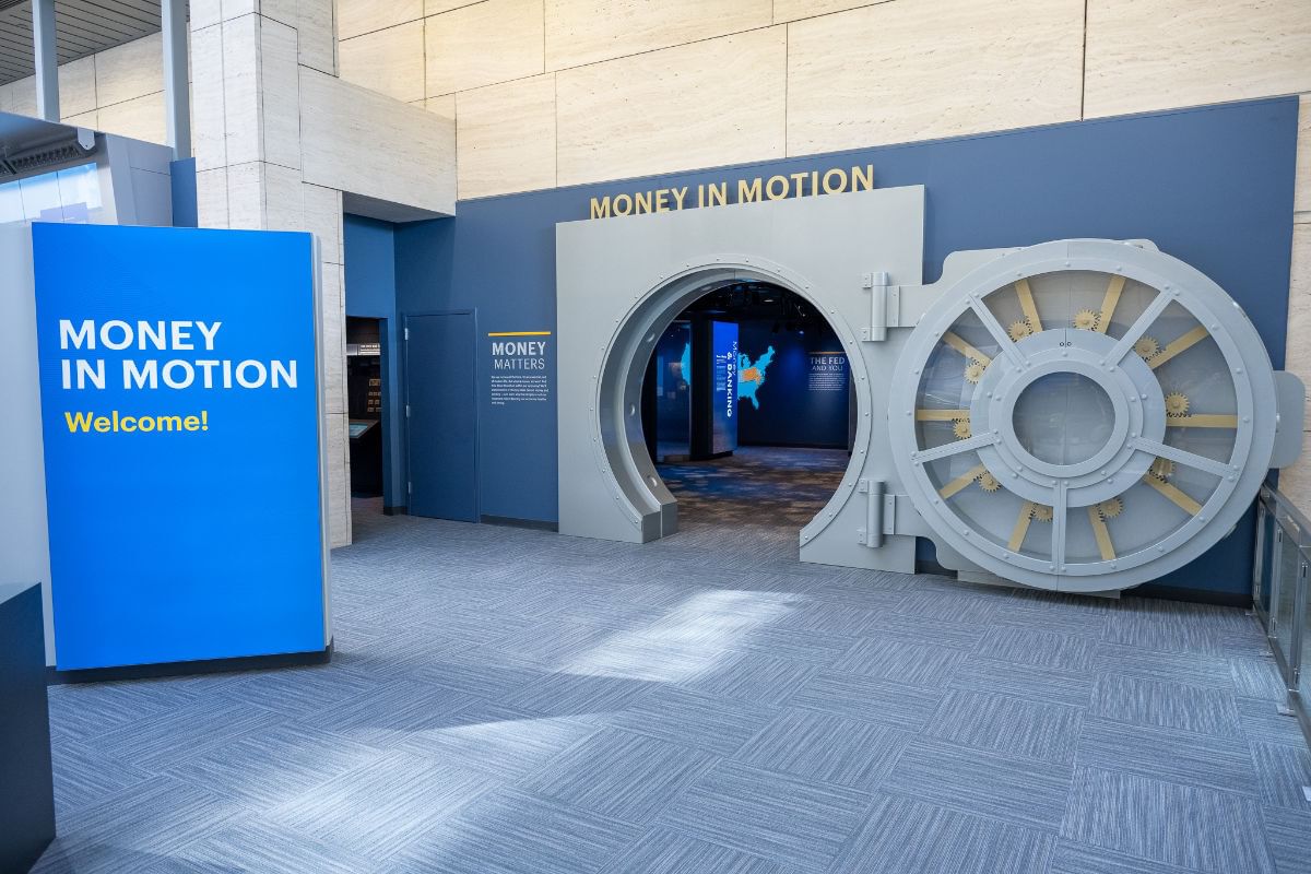 Exhibition entrance view with blue welcome signage and an open door styled like a huge gray vault.